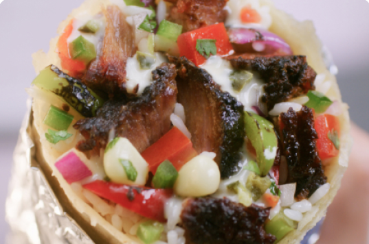 A close up point of view photo of a Smoked Brisket Burrito wrapped in foil and filled with white rice, fajita veggies, hand-chopped Smoked Brisket, fresh tomato salsa, roasted chili corn salsa, and sour cream.